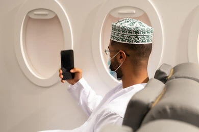 استخدام جهاز تقني حديث, الدردشة مع الأهل والأصدقاء، تصفح مواقع التواصل الاجتماعي, دمج التكنولوجيا المتطورة بالحياة اليومية، صورة من الجانب لرجل عربي خليجي عماني يرتدي الدشداشة والكمة يرتدي قناع حماية كورونا يجلس داخل الطائرة فى رحله جوية يستخدم الهاتف المحمول, مفهوم السياحة و السفر