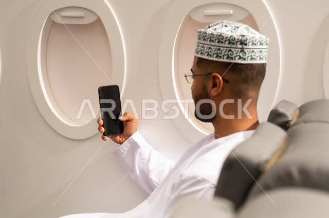 تصفح مواقع التواصل الاجتماعي, الدردشة مع الأهل والأصدقاء، استخدام جهاز تقني حديث, دمج التكنولوجيا المتطورة بالحياة اليومية، صورة من الجانب لرجل عربي خليجي عماني يرتدي الدشداشة والكمة يجلس داخل الطائرة فى رحله جوية يستخدم الهاتف المحمول, مفهوم السياحة و السفر