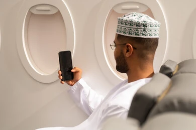 تصفح مواقع التواصل الاجتماعي, الدردشة مع الأهل والأصدقاء، استخدام جهاز تقني حديث, دمج التكنولوجيا المتطورة بالحياة اليومية، صورة من الجانب لرجل عربي خليجي عماني يرتدي الدشداشة والكمة يجلس داخل الطائرة فى رحله جوية يستخدم الهاتف المحمول, مفهوم السياحة و السفر