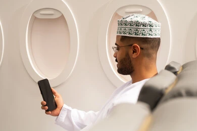 الدردشة مع الأهل والأصدقاء، تصفح مواقع التواصل الاجتماعي، استخدام جهاز تقني حديث, دمج التكنولوجيا المتطورة بالحياة اليومية، صورة من الجانب لرجل عربي خليجي عماني يرتدي الدشداشة والكمة يجلس داخل الطائرة فى رحله جوية يستخدم الهاتف المحمول, مفهوم السياحة و السفر