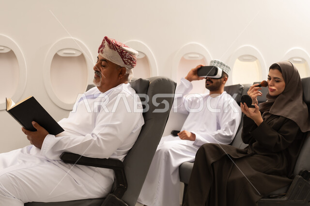 Entertainment and leisure during free time, the concept of tourism and travel, an Omani Gulf Arab family wearing traditional attire sitting inside an airplane on a flight, a Gulf woman applying makeup, the concept of tourism and travel.