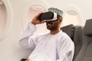 المتعة و التسلية بالمشاهدة عبر نظارة الواقع الافتراضي, مفهوم الاندماج ومحاكاة العالم الخيالي, شاب عربي خليجي عماني يرتدي الدشداشة و الكمة يجلس داخل الطائرة فى رجله جوية ينظر من خلال نظارات ثلاثية الأبعاد، ايماءات وجه تدل على الفرح والسرور, مفهوم السياحة و السفر