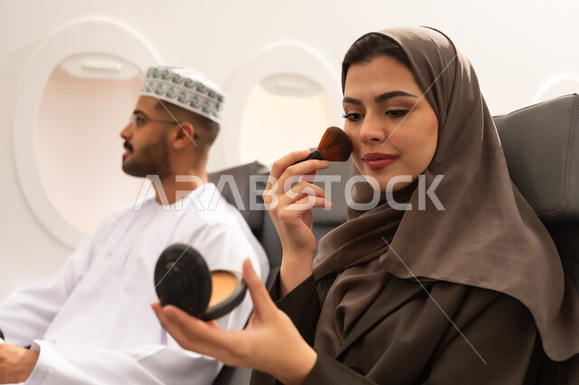 الاهتمام بالأناقة والشكل الخارجي, زوجان عربيان خليجيان عمانيان يرتديان الزي التقليدي يجلسان داخل الطائرة فى رحله جوية , امراة خليجيه تمسك بيدها الفرشاة و تقوم بوضع المكياج , الاستمتاع بقضاء رحله صيفية ممتعه