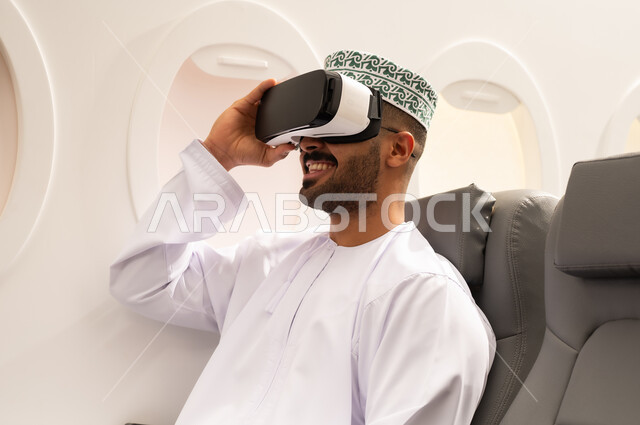 مفهوم الاندماج ومحاكاة العالم الخيالي، المتعة و التسلية بالمشاهدة عبر نظارة الواقع الافتراضي, شاب عربي خليجي عماني يرتدي الدشداشة و الكمة يجلس داخل الطائرة فى رجله جوية ينظر من خلال نظارات ثلاثية الأبعاد، ايماءات وجه تدل على الفرح والسرور, مفهوم السياحة و السفر