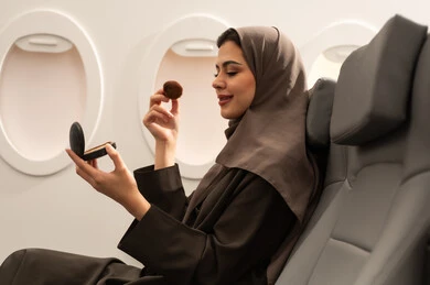 الحرص على الأناقة والاعتناء بالشكل, استخدام المساحيق التجميلية, مفهوم التزين والتجمل, صورة من الجانب لامرأة عربية خليجية عمانية محجبة ترتدي العباءة  تجلس داخل الطائرة فى رحله جوية تستعمل فرشاة أحمر الخدود, شابة سعودية تضع المكياج, مفهوم السياحة و السفر