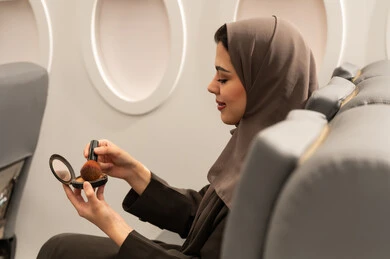 شابة سعودية تضع المكياج, الاهتمام والعناية بالمظهر الخارجي, استخدام المساحيق التجميلية, مفهوم التزين والتجمل, صورة من الجانب لامرأة عربية خليجية عمانية محجبة ترتدي العباءة  تجلس داخل الطائرة فى رحله جوية تستعمل فرشاة أحمر الخدود, مفهوم السياحة و السفر