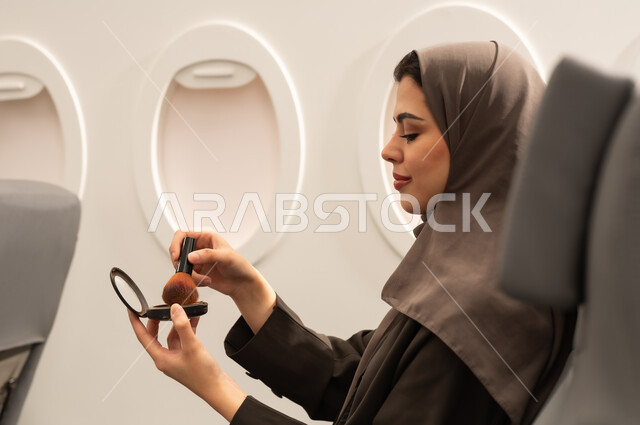مفهوم التزين والتجمل, الاهتمام والعناية بالمظهر الخارجي, استخدام المساحيق التجميلية, صورة من الجانب لامرأة عربية خليجية عمانية محجبة ترتدي العباءة  تجلس داخل الطائرة فى رحله جوية تستعمل فرشاة أحمر الخدود, شابة سعودية تضع المكياج, مفهوم السياحة و السفر