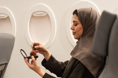 The concept of adornment and beautification, attention and care for the external appearance, the use of cosmetics, a side view of a veiled Gulf Arab Omani woman wearing an abaya sitting inside an airplane on a flight using a blush brush, a young Saudi woman applying makeup, the concept of tourism and travel.