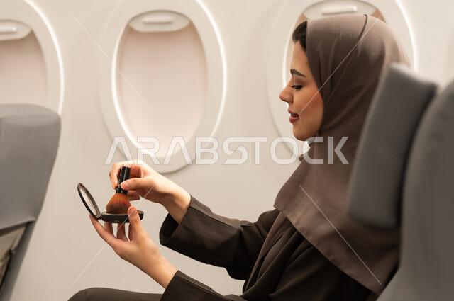 استخدام المساحيق التجميلية, الاهتمام والعناية بالمظهر الخارجي, مفهوم التزين والتجمل, صورة من الجانب لامرأة عربية خليجية عمانية محجبة ترتدي العباءة  تجلس داخل الطائرة فى رحله جوية تستعمل فرشاة أحمر الخدود, شابة سعودية تضع المكياج, مفهوم السياحة و السفر