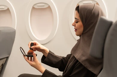 استخدام المساحيق التجميلية, الاهتمام والعناية بالمظهر الخارجي, مفهوم التزين والتجمل, صورة من الجانب لامرأة عربية خليجية عمانية محجبة ترتدي العباءة  تجلس داخل الطائرة فى رحله جوية تستعمل فرشاة أحمر الخدود, شابة سعودية تضع المكياج, مفهوم السياحة و السفر