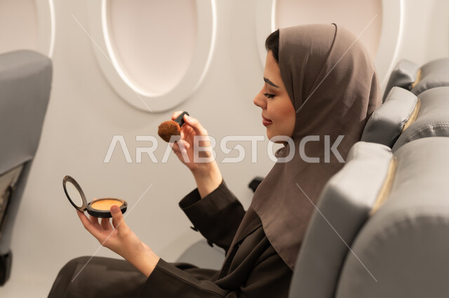 الاهتمام والعناية بالمظهر الخارجي, استخدام المساحيق التجميلية, مفهوم التزين والتجمل, صورة من الجانب لامرأة عربية خليجية عمانية محجبة ترتدي العباءة  تجلس داخل الطائرة فى رحله جوية تستعمل فرشاة أحمر الخدود, شابة سعودية تضع المكياج, مفهوم السياحة و السفر