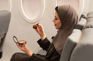 الاهتمام والعناية بالمظهر الخارجي, استخدام المساحيق التجميلية, مفهوم التزين والتجمل, صورة من الجانب لامرأة عربية خليجية عمانية محجبة ترتدي العباءة  تجلس داخل الطائرة فى رحله جوية تستعمل فرشاة أحمر الخدود, شابة سعودية تضع المكياج, مفهوم السياحة و السفر