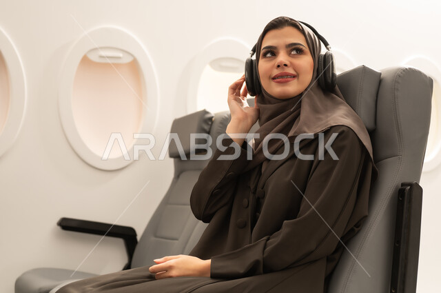استخدام التقنيات الحديثة للتسلية والترفيه, الاستمتاع بسماع الموسيقى المفضلة, قضاء الوقت الممتع, الاندماج مع الألحان الموسيقية, امراة عربيه خليجيه عمانية ترتدي الحجاب و العباءة تجلس داخل الطائرة فى رحله جوية تضع سماعات الرأس الصوتية , مفهوه السياحة و السفر