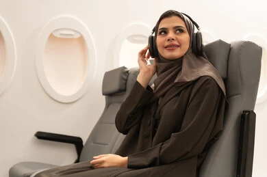 استخدام التقنيات الحديثة للتسلية والترفيه, الاستمتاع بسماع الموسيقى المفضلة, قضاء الوقت الممتع, الاندماج مع الألحان الموسيقية, امراة عربيه خليجيه عمانية ترتدي الحجاب و العباءة تجلس داخل الطائرة فى رحله جوية تضع سماعات الرأس الصوتية , مفهوه السياحة و السفر
