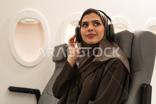 الاستمتاع بسماع الموسيقى المفضلة, استخدام التقنيات الحديثة للتسلية والترفيه, قضاء الوقت الممتع, الاندماج مع الألحان الموسيقية, امراة عربيه خليجيه عمانية ترتدي الحجاب و العباءة تجلس داخل الطائرة فى رحله جوية تضع سماعات الرأس الصوتية , مفهوه السياحة و السفر