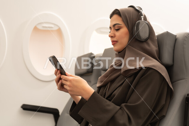 Using technology and modern techniques, communicating with family and friends, watching enjoyable movies and series, following social media sites via mobile phone, a side view of an Arab Gulf Omani woman wearing a hijab and abaya with headphones sitting inside an airplane on a flight holding a mobile phone in her hands, the concept of tourism and travel.