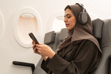 Using technology and modern techniques, communicating with family and friends, watching enjoyable movies and series, following social media sites via mobile phone, a side view of an Arab Gulf Omani woman wearing a hijab and abaya with headphones sitting inside an airplane on a flight holding a mobile phone in her hands, the concept of tourism and travel.