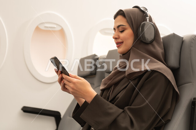التواصل مع الأهل والأصدقاء, مشاهدة الأفلام والمسلسلات الممتعة، متابعة مواقع التواصل الاجتماعي عبر الهاتف المحمول، استخدام التكنولوجيا و التقنيات الحديثة, صورة من الجانب لامراة عربيه خليجيه عمانية ترتدي الحجاب و العباءة و سماعات الرأس تجلس داخل الطائرة فى رحله جويه تحمل  بيديها موبايل، مفهوم السياحه و السفر