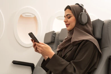 التواصل مع الأهل والأصدقاء, مشاهدة الأفلام والمسلسلات الممتعة، متابعة مواقع التواصل الاجتماعي عبر الهاتف المحمول، استخدام التكنولوجيا و التقنيات الحديثة, صورة من الجانب لامراة عربيه خليجيه عمانية ترتدي الحجاب و العباءة و سماعات الرأس تجلس داخل الطائرة فى رحله جويه تحمل  بيديها موبايل، مفهوم السياحه و السفر