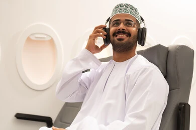 الاستماع للأغاني باسخدام الهيدفون، شاب عربي خليجي عماني يرتدي الدشداشة والكمه يجلس داخل الطائرة فى رحله جوية يضع سماعات الرأس بإيماءات الفرح والسرور، التفاعل والاندماج مع الأنغام الموسيقية, مفهوم السياحة و السفر