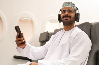 Looking at something, watching enjoyable movies and series, following social media on a mobile phone, communicating with family and friends, using technology and modern techniques, an Arab Gulf Omani man wearing a dishdasha and kumma with headphones sitting inside an airplane on a flight holding a mobile phone, the concept of tourism and travel.