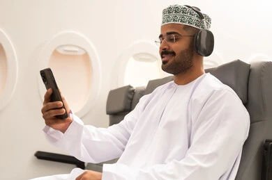 Watching enjoyable movies and series, following social media through mobile phones, communicating with family and friends, using technology and modern techniques, an Arab Gulf Omani man wearing a dishdasha and kumma with headphones sitting inside an airplane on a flight holding a mobile phone, the concept of tourism and travel. Watching enjoyable movies and series, following social media through mobile phones, communicating with family and friends, using technology and modern techniques, an Arab Gulf Omani man wearing a dishdasha and kumma with headphones sitting inside an airplane on a flight holding a mobile phone, the concept of tourism and travel.