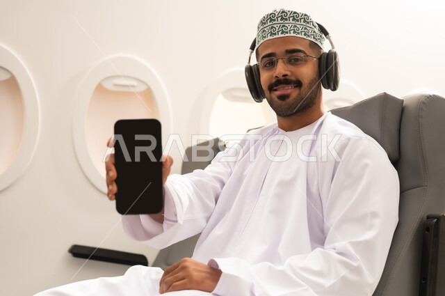 عرض شاشة سوداء فارغة على الجوال، استخدام الوسائل التقنية الحديثة، شاب عربي خليجي عماني يرتدي الدشداشة والكمه و السماعات يجلس داخل الطائرة فى رحله جويه يحمل الهاتف المحمول ينظر  بايماءات السرور, مفهوم السياحه و السفر