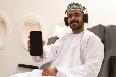 عرض شاشة سوداء فارغة على الجوال، استخدام الوسائل التقنية الحديثة، شاب عربي خليجي عماني يرتدي الدشداشة والكمه و السماعات يجلس داخل الطائرة فى رحله جويه يحمل الهاتف المحمول ينظر  بايماءات السرور, مفهوم السياحه و السفر