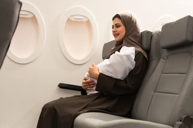 The meaning of motherhood and caring for children, an Arab Gulf Omani mother wearing a hijab and abaya sitting inside the airplane on a flight holding a newborn baby dressed in white cotton clothing, the concept of tourism and travel.
