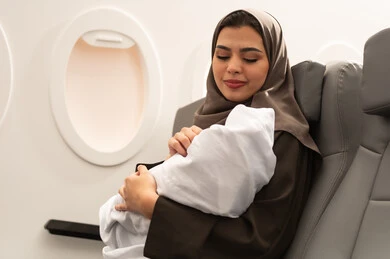 الأمومة وحماية الأطفال ورعايتهم, ام عربية خليجيه عمانية ترتدي الحجاب و العباءة تجلس داخل الطائرة فى رحله جويه تحمل رضيع حديث الولادة يرتدي لباس أبيض قطني, مفهوم السياحه و السفر, ايماءات الحب و الحنان