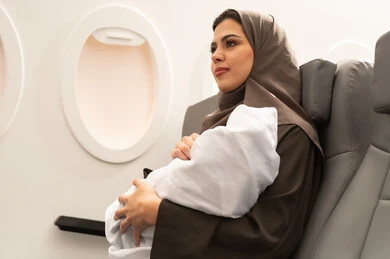 الرعاية والاهتمام بالأبناء, ام عربية خليجيه عمانية ترتدي الحجاب و العباءة تجلس داخل الطائرة فى رحله جويه تحمل رضيع حديث الولادة يرتدي لباس أبيض قطني, مفهوم السياحه و السفر