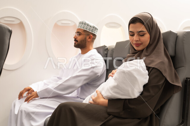 Entertainment and tourist trips during the holiday, the concept of motherhood and child care, an Omani Gulf Arab family wearing traditional attire sitting inside an airplane on a flight, a Gulf mother holding her infant looking at him with gestures of tenderness and love.