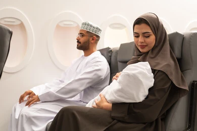 الترفيه والرحلات السياحية في العطلة, مفهوم الامومه والرعايه بالطفل , عائلة عربية خليجيه عمانية ترتدي الزي التقليدي تجلس داخل الطائرة فى رحله جويه , ام خليجيه تحمل طفلها الرضيع تنظر اليه بإيماءات الحنان والحب