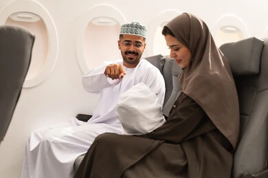 The concept of motherhood and child care, going on recreational trips during vacation, an Arab Gulf Omani family wearing traditional attire sitting inside an airplane on a flight, a Gulf mother holding her infant looking at him with gestures of tenderness and love.