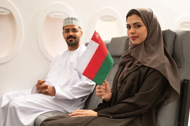 Love of the homeland and belonging to it, taking recreational trips during vacation, the concept of tourism and travel, an Arab Gulf Omani woman wearing a hijab and abaya sitting inside the airplane on a flight with her husband holding the Omani flag in her hand, preparing for an enjoyable trip, gestures of happiness and joy.
