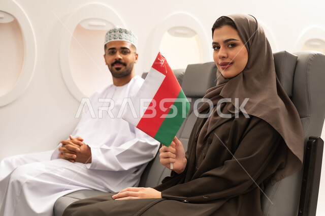 Taking recreational tours during vacation, the concept of tourism and travel, an Arab Gulf Omani woman wearing a hijab and abaya sitting inside the airplane on a flight with her husband holding the Omani flag in her hand, preparing for an enjoyable trip, love for the homeland and belonging to it, gestures of happiness and joy.