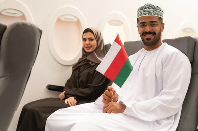 The concept of tourism and travel, taking recreational trips during vacation time, an Arab Gulf Omani man wearing a dishdasha and a kumma sitting inside an airplane on a flight with his wife holding the Omani flag in his hand, preparing for an enjoyable trip, love for the homeland and belonging to it, gestures of happiness and joy.