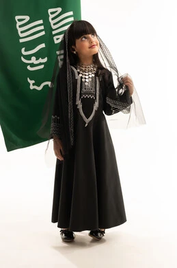 Northern region costume on Saudi National Day, September 23, looking up with gestures of pride in Saudi heritage, the day we began in 1727 AD, portrait of a smiling Saudi Arabian Gulf girl wearing a muhthal and a shayla standing next to the flag of the founding of the first Saudi state on February 22, full body, white background