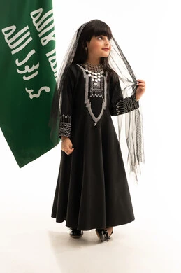 The Day We Began 1727 AD, the costume of the northern region on Saudi National Day, September 23, looking up with gestures of pride in Saudi heritage, a portrait of a smiling Saudi Arabian Gulf girl wearing a muhthal and a shayla standing next to the flag of the founding of the first Saudi state on February 22, full-length body, white background
