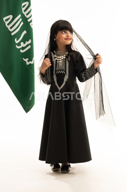 زي المنطقة الشمالية في اليوم الوطني السعودي 23سبتمبر, النظر إلى الأعلى بإيماءات الفخر بالتراث السعودي، يوم بدينا 1727م, بورتريه لفتاة عربية خليجية سعودية مبتسمة ترتدي المحوثل والشيلة تقف بجانب علم تأسيس الدولة السعودية الأولى في 22 فبراير, الجسم بالطول الكامل, خلفية بيضاء
