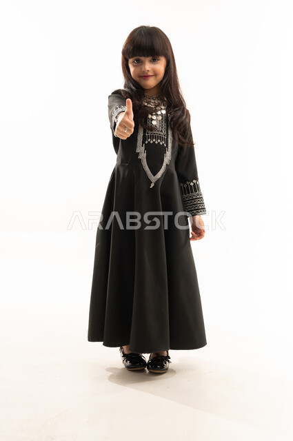 Looking at the camera and giving a thumbs up gesture, wearing the Northern Region costume, the day we started 1727 AD, the anniversary of the founding of the first Saudi state on February 22, portrait of a smiling Saudi Arabian Gulf girl wearing a black muhthal, full body, white background