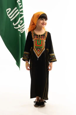 The Day We Began 1727 AD, the anniversary of the founding of the first Saudi state on February 22, wearing the costumes of the southern region on national occasions, a portrait of a Saudi Gulf Arab girl wearing a sheila and a sidrah standing next to the founding flag with gestures of happiness and pride, full-length body, white background