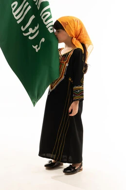 ارتداء أزياء المنطقة الجنوبية في المناسبات الوطنية, التباهي بالانتماء للوطن، يوم بدينا 1727م, ذكرى تأسيس الدولة السعودية الأولى في 22 فبراير, بورتريه لفتاة عربية خليجية سعودية ترتدي الشيلة والسدرة تقوم بتقبيل علم التأسيس بإيماءات الفخر, الجسم بالطول الكامل, خلفية بيضاء