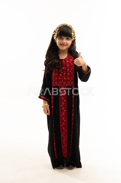 النظر إلى الكاميرا ورفع إصبع الإبهام للأعلى بإيماءات الإعجاب، ارتداء زي المنطقة الشمالية, يوم بدينا 1727م, ذكرى تأسيس الدولة السعودية الأولى في 22 فبراير, بورتريه لفتاة عربية خليجية سعودية مبتسمة ترتدي المحوثل الأسود, الجسم بالطول الكامل, خلفية بيضاء