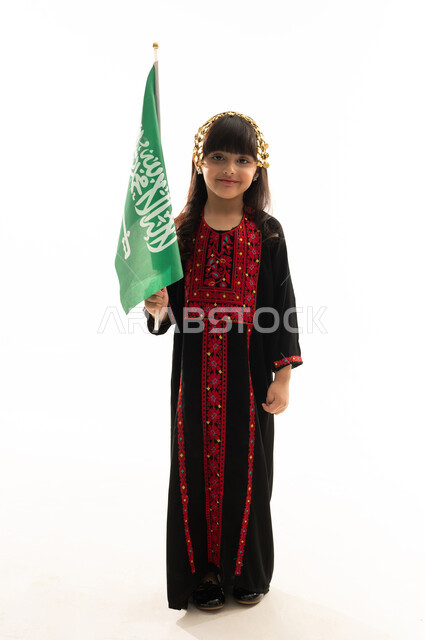 Saudi National Day September 23, Flag Day March 11, wearing the Northern Region costume, the day of our beginnings 1727 AD, the anniversary of the founding of the first Saudi state on February 22, portrait of a smiling Saudi Gulf Arab girl wearing a Mahouthal carrying the Saudi flag with pride and dignity, full body, white background