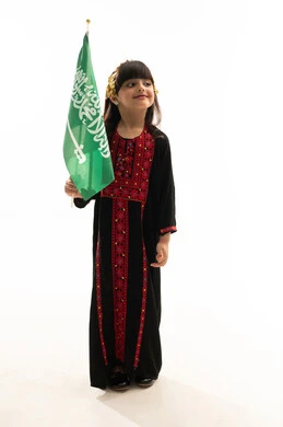 اليوم الوطني السعودي 23سبتمبر، يوم العلم 11مارس، ارتداء زي المنطقة الشمالية, يوم بدينا 1727م, ذكرى تأسيس الدولة السعودية الأولى في 22 فبراير, بورتريه لفتاة عربية خليجية سعودية مبتسمة ترتدي المحوثل تحمل العلم السعودي بفخر وشموخ, الجسم بالطول الكامل, خلفية بيضاء