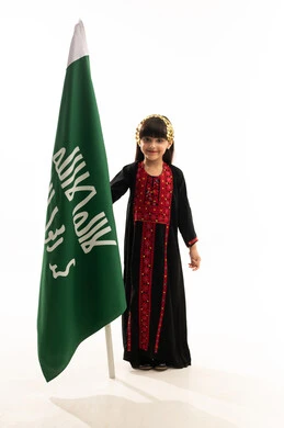 تنشئة الابناء على حب الوطن، يوم بدينا 1727م, زي المنطقة الشمالية في اليوم الوطني السعودي23سبتمبر, النظر إلى الكاميرا بفخر، بورتريه لفتاة عربية خليجية سعودية مبتسمة ترتدي المحوثل تمسك علم تأسيس الدولة السعودية الأولى في 22 فبراير, الجسم بالطول الكامل, خلفية بيضاء