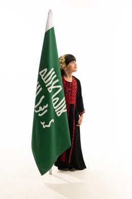 حب الوطنوالانتماء إليه، يوم بدينا 1727م, زي المنطقة الشمالية في المناسبات الوطنية, النظر إلى الأعلى بفخر، بورتريه لفتاة عربية خليجية سعودية مبتسمة ترتدي المحوثل تمسك علم تأسيس الدولة السعودية الأولى في 22 فبراير, الجسم بالطول الكامل, خلفية بيضاء