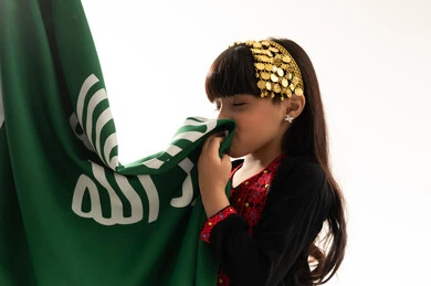 اليوم الوطني السعودي 23سبتمبر، الاعتزاز بالانتماء إليه، يوم بدينا 1727م, زي المنطقة الشمالية في المناسبات الوطنية, النظر إلى الأعلى بفخر، بورتريه مقرب لفتاة عربية خليجية سعودية ترتدي المحوثل تقوم بتقبيل علم تأسيس الدولة السعودية الأولى في 22 فبراير, خلفية بيضاء