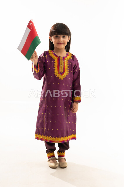 إحياء ذكرى تأسيس الدولة 1775 م، النظر إلى الكاميرا بفخر، رموز وثقافات وطنية ,  حب الوطن والانتماء إليه، بورتريه لفتاة عربية خليجية عمانية ترتدي الزي الشعبي التقليدي تحمل العلم العماني، الاحتفال بذكرى اليوم الوطني العماني 18 نوفمبر، الجسم بالطول الكامل، خلفية بيضاء